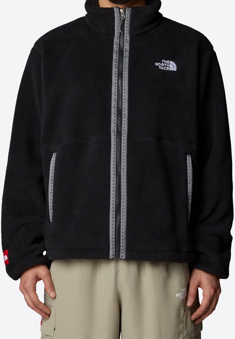 Svart fleecejacka med hög krage, dragkedjestängning, två sidofickor och grå sömmar. Har The North Face-logotypen på bröstet.