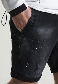 Pantalones cortos de mezclilla negra con detalles desgastados, acentos de salpicaduras de pintura y dobladillos enrollados. El material es suave con un ligero brillo.