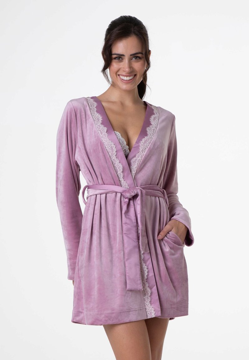 Accappatoio di velluto lavanda con maniche lunghe, scollo a V profondo, bordi in pizzo lungo il colletto, vita legata e tasche laterali. Texture liscia e morbida.