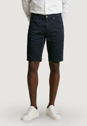 Hombre que lleva pantalones cortos casuales negros hasta la rodilla y zapatillas blancas, de pie con las manos detrás de la espalda sobre un fondo liso.