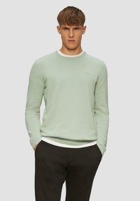 Hellgrüner Strickpullover mit rundem Halsabschluss, versehen mit einem kontrastierenden weißen Kragen. Getragen über einem weißen Hemd und kombiniert mit dunklen Hosen.