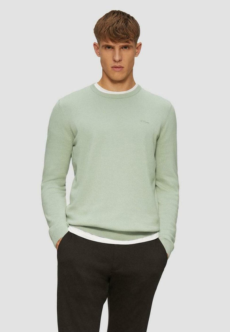 Hellgrüner Strickpullover mit rundem Halsabschluss, versehen mit einem kontrastierenden weißen Kragen. Getragen über einem weißen Hemd und kombiniert mit dunklen Hosen.