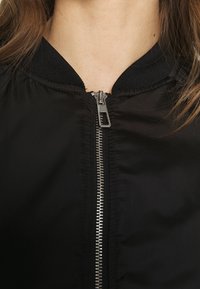 Czarna kurtka bombowa z zamkiem z przodu, prążkowanym kołnierzem i gładkim, satynowym wykończeniem. Zawiera srebrny metalowy detal zamka.