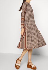 Hnědé šaty s vzorem, které mají kulatý výstřih, dlouhé rukávy a rozšířenou sukni. Doplněné hnědými platformovými sandály se pásky.