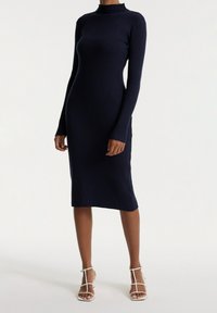 Robe en tricot côtelé marine avec un col haut et des manches longues, présentant une silhouette ajustée qui tombe sous le genou. Accentuée avec des sandales blanches à lanières.