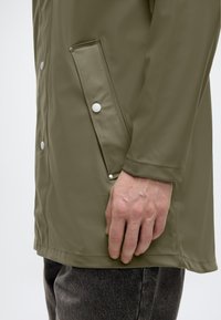 Waterdichte jas in olijfgroen met een gladde textuur, drukknopen en een zijzak met een afgeronde klep. Gedragen over een donkere spijkerbroek.