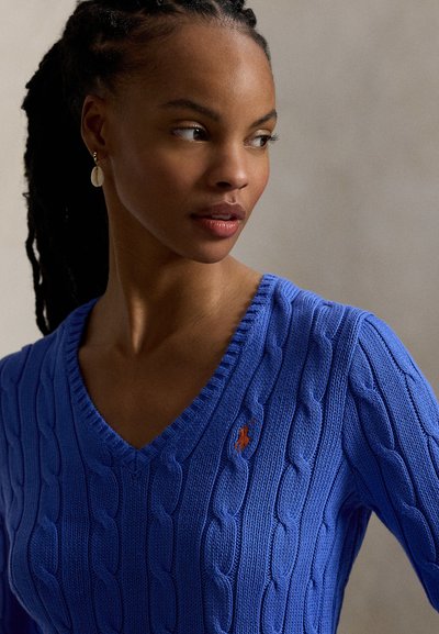 Μπλε πουλόβερ με πλέξη καλωδίου και V-λαιμόκοψη. Διαθέτει μικρό κόκκινο λογότυπο. Η υφή φαίνεται μαλακή και με ριγέ λεπτομέρειες, προσφέροντας μια κομψή εμφάνιση.