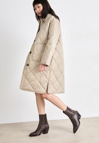 Manteau matelassé beige avec une fermeture à boutons et des poches latérales, associé à des bottines marron à bout pointu et à talon effilé.