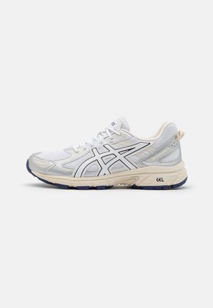 Chaussure de course blanche et grise avec des bandes noires du logo, semelle rembourrée indiquée "GEL" et semelle bleu marine, vue de côté sur un fond blanc.