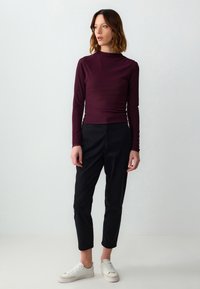 Langärmliges, eng anliegendes Oberteil in tiefem Burgunderrot, kombiniert mit hochgeschnittenen schwarzen Hosen. Weiße Sneaker runden das Outfit ab. Glatte Textur.