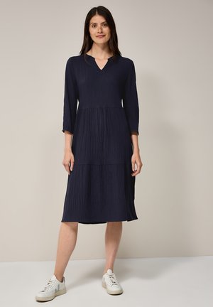 Robe marine longueur genou en tissu texturé avec un décolleté en V et des manches trois-quarts, associée à des baskets blanches pour un look décontracté.