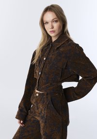 Brown and navy patterned cropped jacket with a collar, front buttons, and two chest pockets, paired with high-waisted trousers.