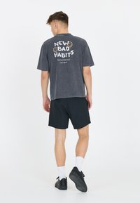 Nuori mies kävelee pois päällään haalistunut musta t-paita, jossa on teksti "NEW BAD HABITS" ja hymiöitä, mustat shortsit, valkoiset sukat ja mustat kengät.