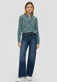 Blusa floreale in turchese, verde e bianco, abbinata a jeans scuri a vita alta. Il modello indossa sneakers bianche spesse, appoggiato su uno sfondo neutro.