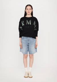 Čierna mikina s zeleným nápisom "ARMANI EXCHANGE", v kombinácii s svetlomodrými džínsovými šortkami s roztrhnutým okrajom, bielymi ponožkami a béžovými teniskami.