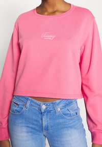 Persona vestida con un sweatshirt corto rosa con el logo de "Tommy Jeans" y jeans de denim azul claro, de pie contra un fondo liso.