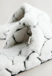 Hvid, fluffy tøffel med en blød tekstur, med et sort geometrisk mønster og en lille mærkat med teksten "NEXT" på indersiden.