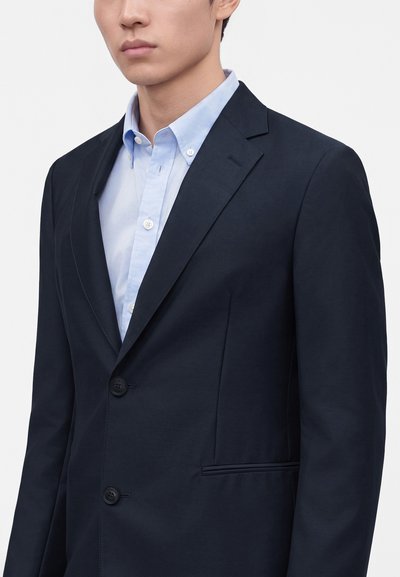 Homme portant un blazer bleu marine foncé sur une chemise bleu clair à boutons, sur un fond blanc uni.