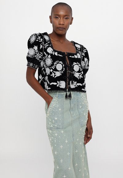 Μαύρη κεντητή τοπ με φουσκωτά μανίκια και άνοιγμα στον μπροστινό μέρος, συνδυασμένο με μια ανοιχτό μπλε τζην φούστα διακοσμημένη με λευκές χάντρες.