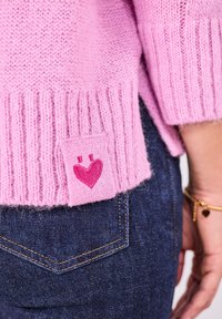 Rosa gestrickter Pullover mit Rippenbund, der ein kleines Herz-Patch aufweist. Getragen mit dunkelblauen Jeans, die eine Stichdetail auf der Tasche zeigen.