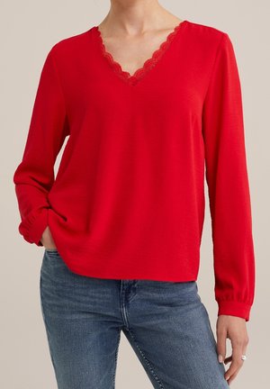 Vrouw die een felrode blouse met lange mouwen en een V-hals met kanten rand draagt, gecombineerd met een blauwe spijkerbroek, staand met één hand in haar zak.