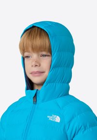 Μπλε επενδυμένο μπουφάν με κουκούλα, με υφή καπιτονέ και κλείσιμο με φερμουάρ μπροστά. Λογότυπο εμφανίζεται στην αριστερή πλευρά του στήθους, με λείο φινίρισμα.