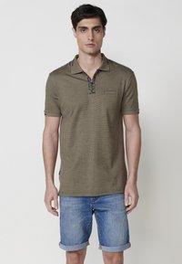 Camisa polo de color verde oliva con tejido texturizado, mangas cortas, un cuello con media cremallera y un pequeño bolsillo en el pecho. Combinada con pantalones cortos de denim enrollados.