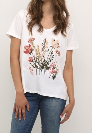Femme portant une chemise blanche à manches courtes avec un motif floral multicolore et un jean bleu, vue du cou aux hanches sur un fond uni.