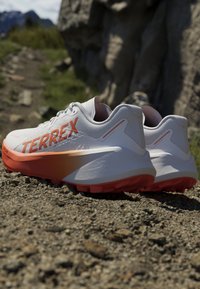 Par hvide og orange Terrex trail løbesko på stenrig udendørs jord nær en stenvæg med sløret naturlandskab i baggrunden.
