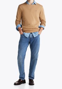 Homme portant un pull beige par-dessus une chemise en denim bleu, un jean bleu et des mocassins marron foncé, debout les mains dans les poches sur fond blanc.
