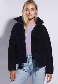 Sort pufferjakke med høj krave, der har lynlåslukning og quiltet design. Båret over en lys lilla top med denimjeans.