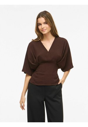 Femme portant un haut marron à col en V avec des manches larges et une taille ajustée, associé à un pantalon noir ample, debout avec une main dans la poche.