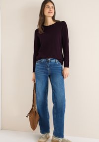 Lange mouw, donkerpaarse trui gecombineerd met blauwe rechte jeans. Het model houdt een bruine suède schoudertas vast en draagt lichte sneakers.