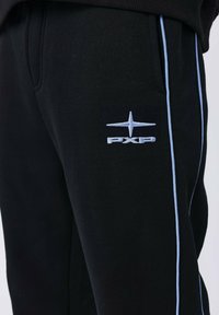 Primer plano de pantalones de chándal negros con ribetes azul claro y un logo bordado de estrella de cuatro puntas en azul claro sobre las letras "PXP" en el muslo.