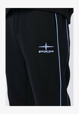 Primer plano de pantalones de chándal negros con ribetes azul claro y un logo bordado de estrella de cuatro puntas en azul claro sobre las letras "PXP" en el muslo.