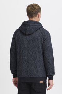 Young man seen from behind wearing a dark gray hooded sweatshirt and black pants against a plain white background.