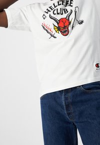 Camiseta blanca de algodón con un estampado gráfico en rojo y negro que muestra la cara de un demonio y llamas, combinada con jeans de mezclilla azul.