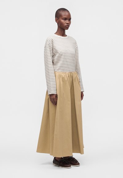 Femme aux cheveux courts portant une longue jupe beige, un haut à manches longues rayé et des chaussures marron foncé, debout devant un fond clair uni.
