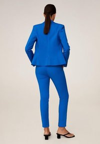 Mujer con cabello oscuro en una coleta, vestida con un traje entallado de color azul brillante y tacones abiertos negros, de pie con las manos en los bolsillos, mirando hacia otro lado.