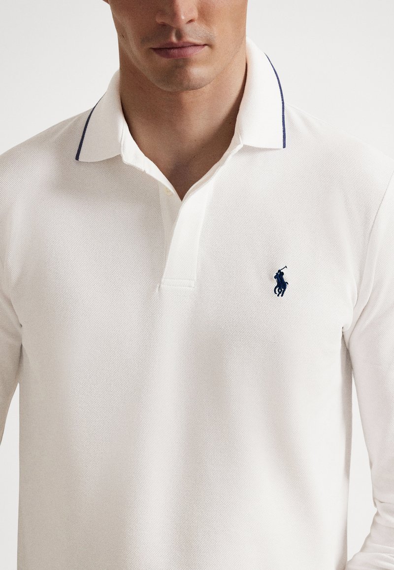Homme portant un polo à manches longues blanc avec des bordures bleu marine sur le col et un petit logo brodé bleu marine sur la poitrine.