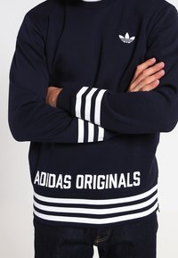 Sudadera azul marino con rayas blancas en los puños y el dobladillo inferior. Presenta un pequeño logo blanco en el pecho. El texto dice "ADIDAS ORIGINALS."