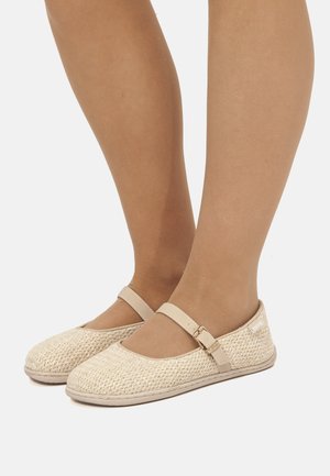 Piedi che indossano scarpe piatte stile Mary Jane intrecciate beige con cinturini regolabili con fibbia su uno sfondo chiaro semplice.