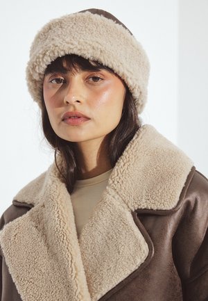 Naine, kes kannab pruuni nahktagi beeži voodriga ja sobivat karvaga vooderdatud mütsi, seisab veidi vasakule ja tal on neutraalne ilme.