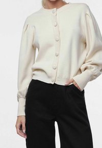 Cárdigan de manga corta en color crema con cuello y puños acanalados, que presenta cinco botones redondos en la parte delantera. Combinado con pantalones negros.