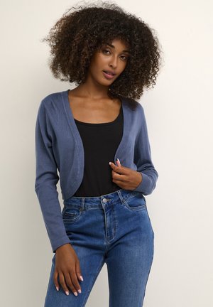 Blue cropped cardigan made of soft material with a v-neck design and long sleeves, worn over a black top and paired with blue jeans.