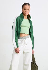 Veste zippée verte et blanche avec un design cropped, haut à côtes vert structuré, pantalon de survêtement blanc et sac vert foncé.