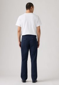 Homme debout, de dos, portant un t-shirt blanc à manches courtes, un pantalon bleu marine avec des poches arrière, et des chaussures noires sur un fond uni.