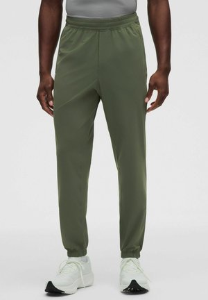 Uomo che indossa pantaloni jogger verde oliva con vita e polsini elastici, abbinati a scarpe da ginnastica bianche e una maglietta grigia.