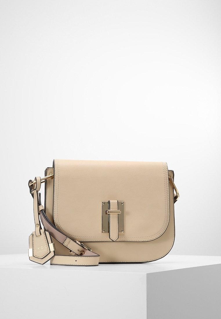 Beżowa torba na ramię ze skóry o wyrazistej, usztywnionej formie, z elementami w odcieniu złota, przednią klapą oraz regulowanym paskiem. Posiada detal z metką.