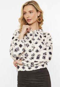 Krämfärgad blus med svarta blommönster, elastiska ärmslut och en rynkad halsringning med dragsko. Den strukturerade tyget ger en lätt känsla.